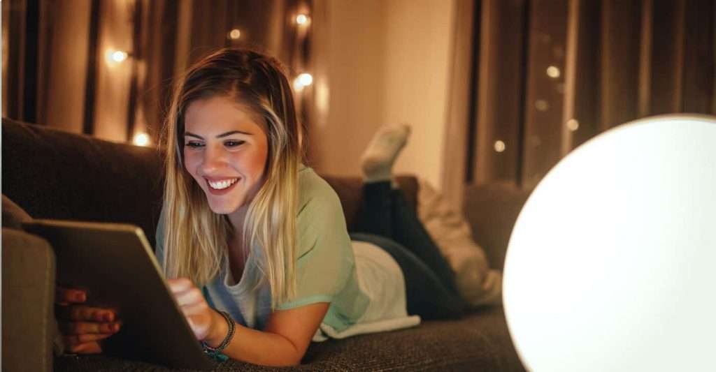 En ung kvinde ligger på en sofa og bruger en tablet. Baggrunden er oplyst med små lys, hvilket skaber en hyggelig atmosfære.