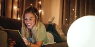 En ung kvinde ligger på en sofa og bruger en tablet. Baggrunden er oplyst med små lys, hvilket skaber en hyggelig atmosfære.