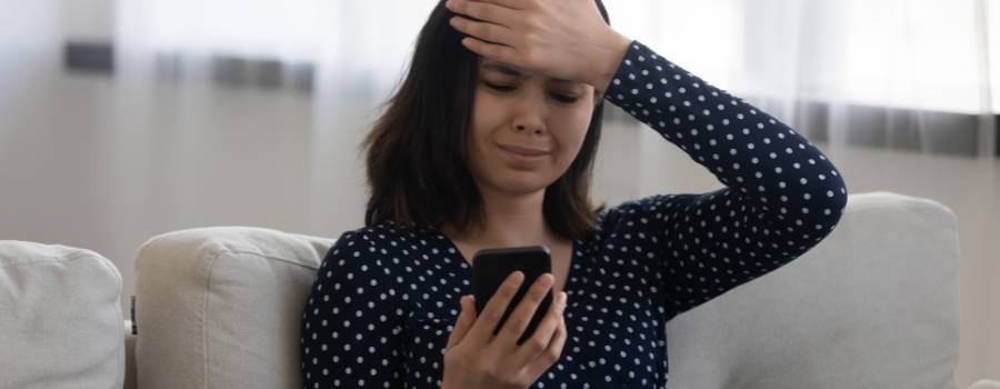 En frustreret asiatisk pige sidder i sofaen med en smartphone og holder sig for panden