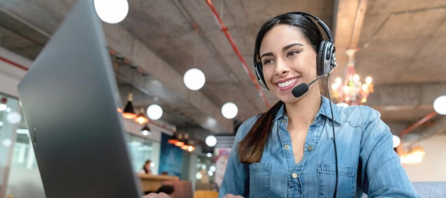 Sådan bliver du virtuel assistent – din guide til et fleksibelt arbejdsliv online 1 En smilende ung kvinde med langt mørkt hår sidder foran en bærbar computer i et åbent kontorlandskab. Hun bærer et headset med en mikrofon, hvilket indikerer, at hun taler eller lytter. Hendes blik er rettet mod skærmen, og bag hende ses et moderne kontormiljø med industrielt loft og belysning.