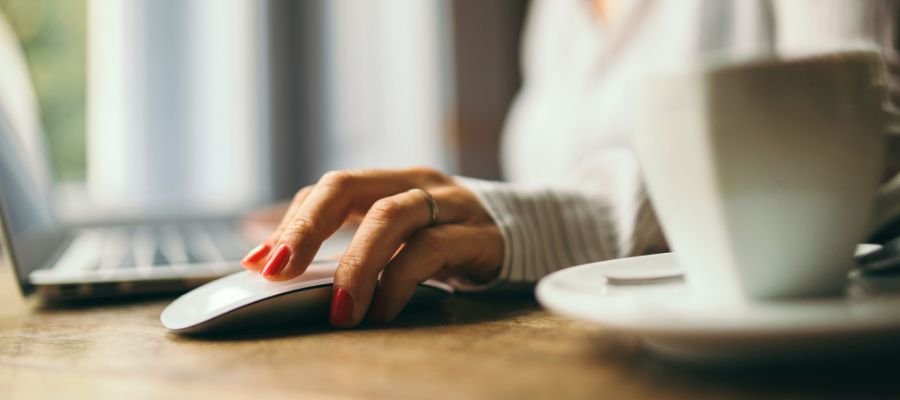 Sådan bliver du virtuel assistent – din guide til et fleksibelt arbejdsliv online 2 Et nærbillede af en hånd med rødlakerede negle, der hviler på en hvid computermus. I baggrunden ses sløret en bærbar computer og en hvid kaffekop på en underkop. Bordet ser ud til at være af træ.