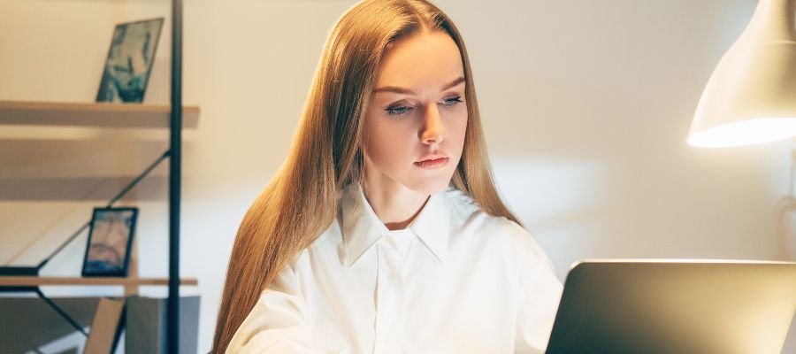 Sådan bliver du virtuel assistent – din guide til et fleksibelt arbejdsliv online 3 En kvinde med langt, lyst hår sidder foran en bærbar computer i et svagt oplyst rum. Hun er klædt i en hvid skjorte og ser koncentreret ned på skærmen. I baggrunden ses en hylde med nogle genstande og en tændt bordlampe.