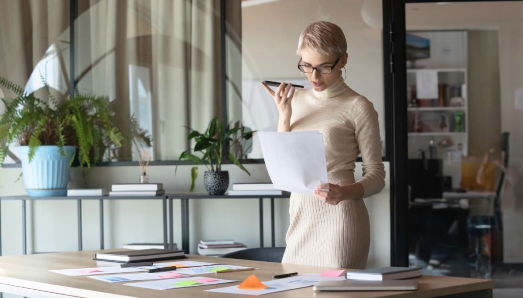 virtuel-assistent-top En kvinde med kort, lyst hår og briller står i et moderne kontormiljø. Hun holder en mobiltelefon op til munden, som om hun taler i den, og holder samtidig et stykke papir i den anden hånd. Rundt om hende er der planter og kontormøbler, herunder et stort skrivebord med papirer og en bærbar computer.