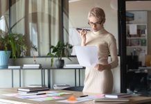 Sådan bliver du virtuel assistent – din guide til et fleksibelt arbejdsliv online En kvinde med kort, lyst hår og briller står i et moderne kontormiljø. Hun holder en mobiltelefon op til munden, som om hun taler i den, og holder samtidig et stykke papir i den anden hånd. Rundt om hende er der planter og kontormøbler, herunder et stort skrivebord med papirer og en bærbar computer.