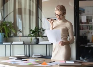 En kvinde med kort, lyst hår og briller står i et moderne kontormiljø. Hun holder en mobiltelefon op til munden, som om hun taler i den, og holder samtidig et stykke papir i den anden hånd. Rundt om hende er der planter og kontormøbler, herunder et stort skrivebord med papirer og en bærbar computer.