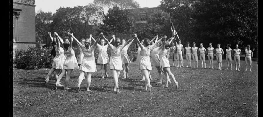 Fotografiet viser en gruppe mennesker, der udfører synkroniserede øvelser eller dansetrin udendørs. De er klædt i lyst sportstøj. Danmark, 1923.