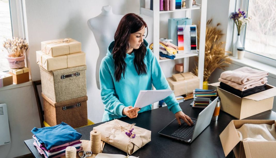 small-business En kvinde arbejder ved en computer i et lille kreativt værksted, omgivet af emballerede varer og stof.