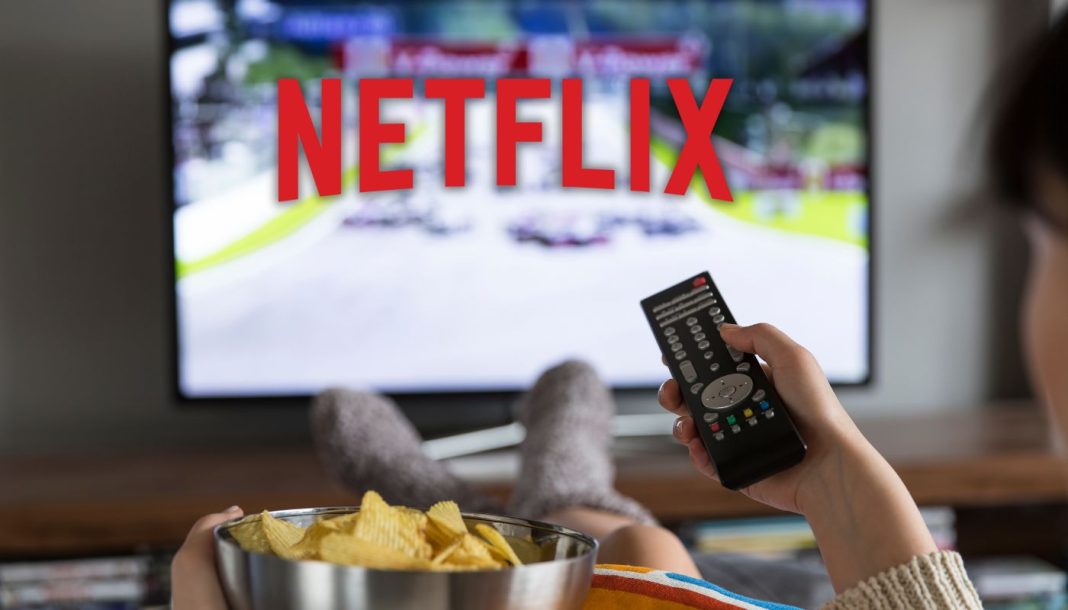En kvinde sidder foran fjernsynet med Netflix-logoet på skærmen, holder en fjernbetjening i den ene hånd og en skål chips i den anden.