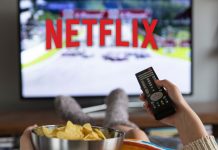 En kvinde sidder foran fjernsynet med Netflix-logoet på skærmen, holder en fjernbetjening i den ene hånd og en skål chips i den anden.