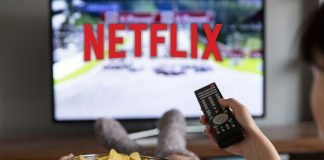 En kvinde sidder foran fjernsynet med Netflix-logoet på skærmen, holder en fjernbetjening i den ene hånd og en skål chips i den anden.