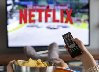 En kvinde sidder foran fjernsynet med Netflix-logoet på skærmen, holder en fjernbetjening i den ene hånd og en skål chips i den anden.