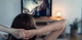 En kvinde sidder på en sofa med hænderne bag hovedet og ser fjernsyn.