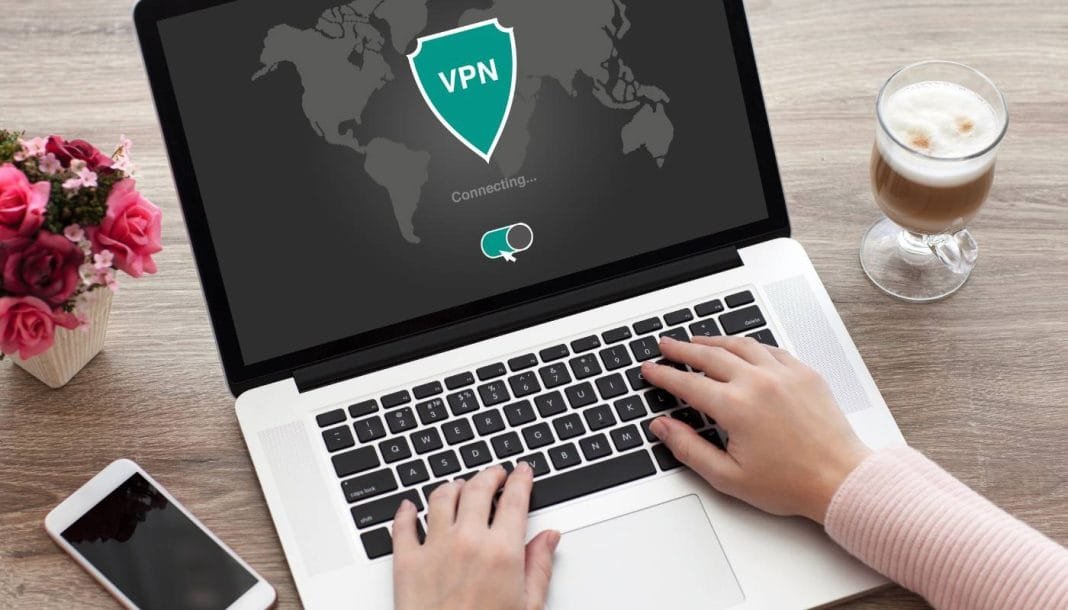 En person bruger en bærbar computer, hvor skærmen viser, at en VPN-forbindelse er ved at blive oprettet. På bordet ved siden af computeren er der en smartphone, en kop kaffe og en vase med lyserøde roser
