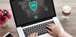 En person bruger en bærbar computer, hvor skærmen viser, at en VPN-forbindelse er ved at blive oprettet. På bordet ved siden af computeren er der en smartphone, en kop kaffe og en vase med lyserøde roser