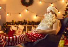 En mand klædt ud som julemanden, der sidder afslappet i en stol. Han har en hvid tanktop, røde og hvide stribede sokker og røde shorts med hvide prikker.