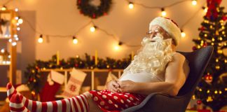 En mand klædt ud som julemanden, der sidder afslappet i en stol. Han har en hvid tanktop, røde og hvide stribede sokker og røde shorts med hvide prikker.