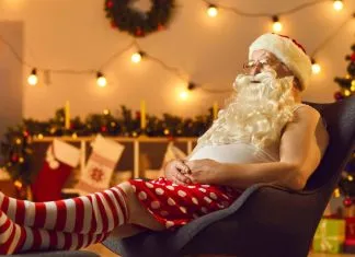 De 17 bedste julefilm at se på Disney+ En mand klædt ud som julemanden, der sidder afslappet i en stol. Han har en hvid tanktop, røde og hvide stribede sokker og røde shorts med hvide prikker.