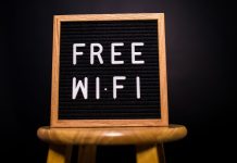 Træramme med teksten 'Free Wi-Fi' skrevet med hvide bogstaver på en sort baggrund, placeret på en træskammel.