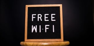 Træramme med teksten 'Free Wi-Fi' skrevet med hvide bogstaver på en sort baggrund, placeret på en træskammel.