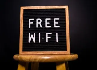 Træramme med teksten 'Free Wi-Fi' skrevet med hvide bogstaver på en sort baggrund, placeret på en træskammel.