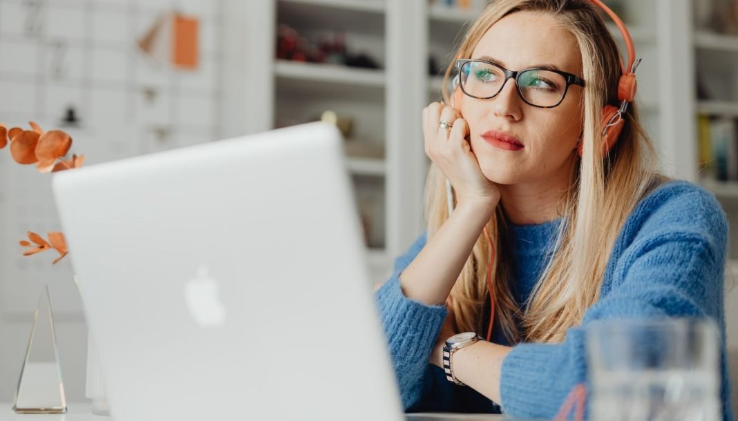 En kvinde med langt lyst hår sidder ved et skrivebord og arbejder på en bærbar computer. Hun har orange hovedtelefoner på og er iført en blå sweater.