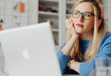 En kvinde med langt lyst hår sidder ved et skrivebord og arbejder på en bærbar computer. Hun har orange hovedtelefoner på og er iført en blå sweater.