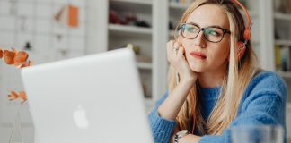 En kvinde med langt lyst hår sidder ved et skrivebord og arbejder på en bærbar computer. Hun har orange hovedtelefoner på og er iført en blå sweater.