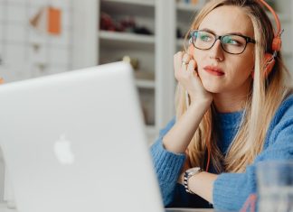 En kvinde med langt lyst hår sidder ved et skrivebord og arbejder på en bærbar computer. Hun har orange hovedtelefoner på og er iført en blå sweater.