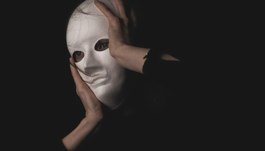 En person holder en hvid maske foran ansigtet i en mørk og mystisk atmosfære.