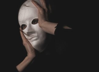 En person holder en hvid maske foran ansigtet i en mørk og mystisk atmosfære.