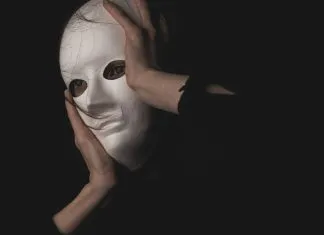 En person holder en hvid maske foran ansigtet i en mørk og mystisk atmosfære.