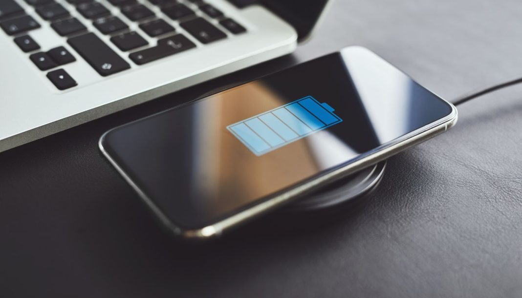 Billedet viser en smartphone, der oplades trådløst ved hjælp af en opladningsplade. Telefonen ligger på opladningspladen, og skærmen viser et batteriikon, der indikerer, at enheden oplades. Ved siden af telefonen er der en bærbar computer, hvoraf en del af tastaturet er synligt.