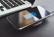 Billedet viser en smartphone, der oplades trådløst ved hjælp af en opladningsplade. Telefonen ligger på opladningspladen, og skærmen viser et batteriikon, der indikerer, at enheden oplades. Ved siden af telefonen er der en bærbar computer, hvoraf en del af tastaturet er synligt.