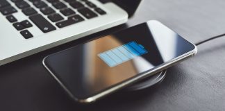 Billedet viser en smartphone, der oplades trådløst ved hjælp af en opladningsplade. Telefonen ligger på opladningspladen, og skærmen viser et batteriikon, der indikerer, at enheden oplades. Ved siden af telefonen er der en bærbar computer, hvoraf en del af tastaturet er synligt.