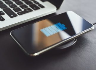 Billedet viser en smartphone, der oplades trådløst ved hjælp af en opladningsplade. Telefonen ligger på opladningspladen, og skærmen viser et batteriikon, der indikerer, at enheden oplades. Ved siden af telefonen er der en bærbar computer, hvoraf en del af tastaturet er synligt.