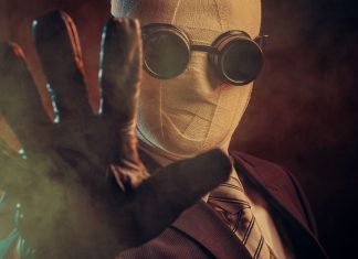 En person iført et jakkesæt, slips og specialiserede mørke briller, med ansigtet dækket af bandager, der rækker en handsket hånd frem mod kameraet. Baggrunden er mørk og tåget, hvilket skaber en mystisk og dramatisk atmosfære.