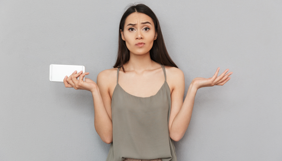 En kvinde med langt, mørkt hår står foran en grå væg. Hun holder en hvid smartphone i venstre hånd og har begge arme løftet med håndfladerne opad, som om hun er forvirret eller spørgende. Hun bærer en grå top med tynde stropper.