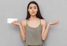 En kvinde med langt, mørkt hår står foran en grå væg. Hun holder en hvid smartphone i venstre hånd og har begge arme løftet med håndfladerne opad, som om hun er forvirret eller spørgende. Hun bærer en grå top med tynde stropper.