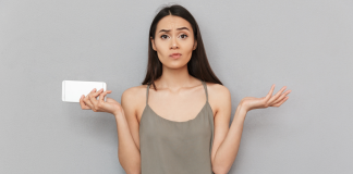 En kvinde med langt, mørkt hår står foran en grå væg. Hun holder en hvid smartphone i venstre hånd og har begge arme løftet med håndfladerne opad, som om hun er forvirret eller spørgende. Hun bærer en grå top med tynde stropper.