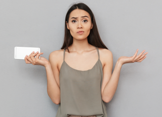 En kvinde med langt, mørkt hår står foran en grå væg. Hun holder en hvid smartphone i venstre hånd og har begge arme løftet med håndfladerne opad, som om hun er forvirret eller spørgende. Hun bærer en grå top med tynde stropper.