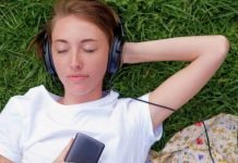 En teenagepige ligger på græsset med hovedtelefoner på og holder en mobiltelefon i hånden. Hun har en hvid t-shirt på, og der er et stykke stof med blomsterprint ved siden af.