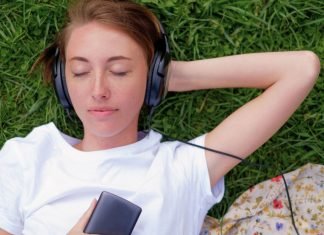 En teenagepige ligger på græsset med hovedtelefoner på og holder en mobiltelefon i hånden. Hun har en hvid t-shirt på, og der er et stykke stof med blomsterprint ved siden af.