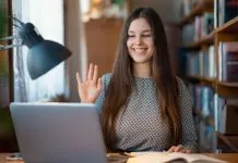 6 tips til at beskytte dit webcam – alle bør kende dem En ung kvinde sidder ved et skrivebord med en bærbar computer og en åben bog. Hun har langt hår og er iført en mønstret trøje. Hun holder hånden op, som om hun vinker eller deltager i et online møde.