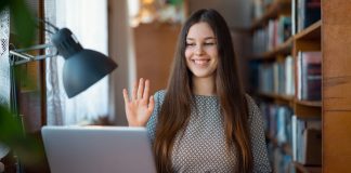 En ung kvinde sidder ved et skrivebord med en bærbar computer og en åben bog. Hun har langt hår og er iført en mønstret trøje. Hun holder hånden op, som om hun vinker eller deltager i et online møde.