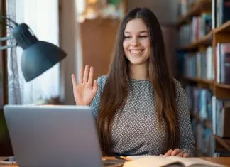 En ung kvinde sidder ved et skrivebord med en bærbar computer og en åben bog. Hun har langt hår og er iført en mønstret trøje. Hun holder hånden op, som om hun vinker eller deltager i et online møde.