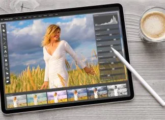 En tablet på et træbord ved siden af en kop kaffe. På tablettens skærm er et foto redigeringsprogram åbent, hvor et billede af en kvinde i en hvid kjole i en mark med høje græsstrå redigeres.