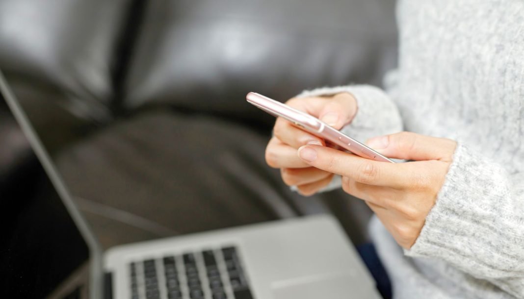 Person bruger en smartphone foran en bærbar computer i sofaen.