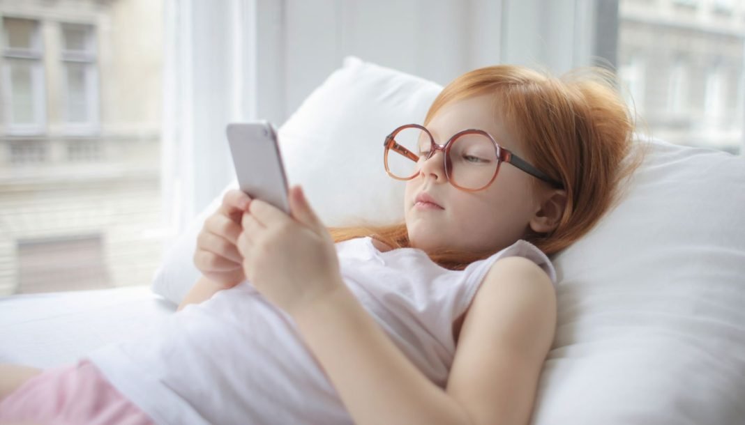 girl-with-smartphone En pige med rødt hår og store briller ligger på en pude og kigger på en smartphone.