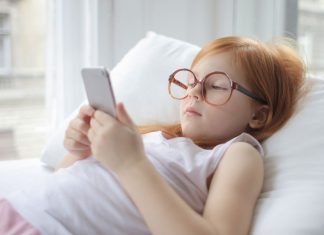 En pige med rødt hår og store briller ligger på en pude og kigger på en smartphone.