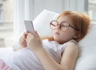 En pige med rødt hår og store briller ligger på en pude og kigger på en smartphone.